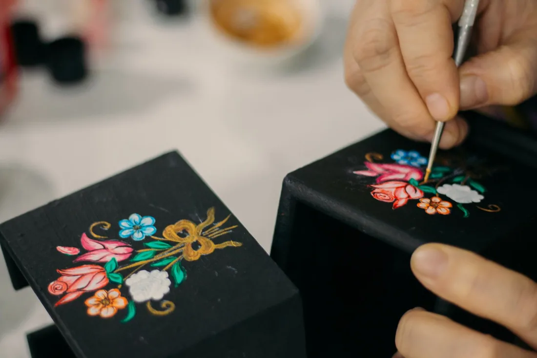 Mano de un artesano pintando un motivo floral con pincel fino sobre una caja laqueada negra